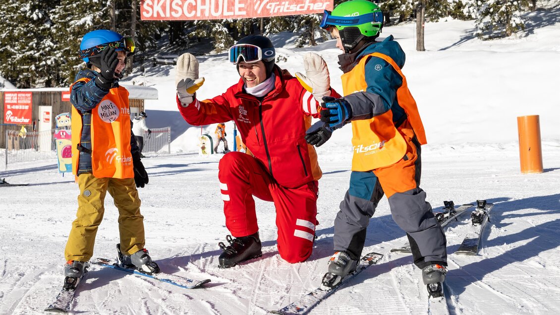 Skilehrerin kniet im Schnee und klatscht mit zwei Kindern ab; im Hintergrund ein Schild der Skischule und weitere Kinder auf der Piste. | © Skischule Tritscher