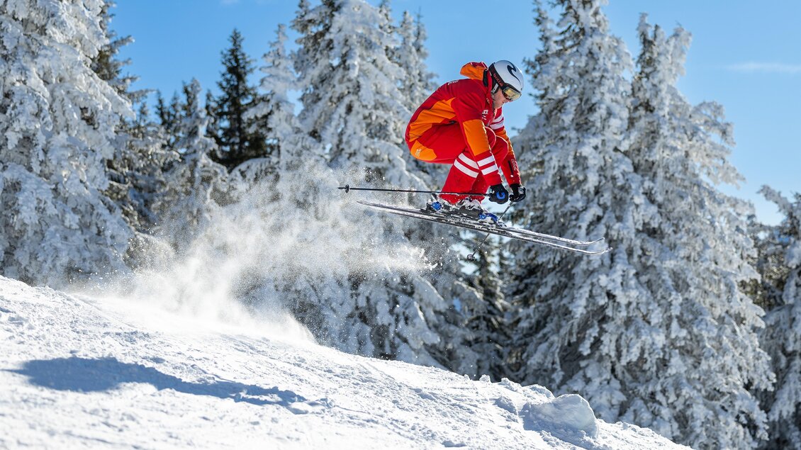 Skilehrer in roter Skischuluniform springt über eine kleine Schneekante; aufgewirbelter Pulverschnee, verschneite Tannen im Hintergrund und klarer blauer Himmel. | © Skischule Tritscher