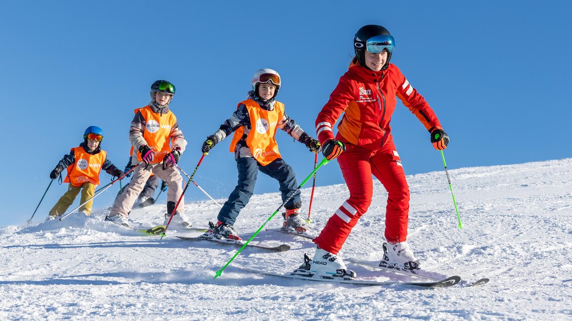 Skilehrerin in roter Skischuluniform fährt vorneweg, dahinter mehrere Kinder mit orangefarbenen Westen beim gemeinsamen Skifahren auf einer sonnigen Piste. | © Skischule Tritscher