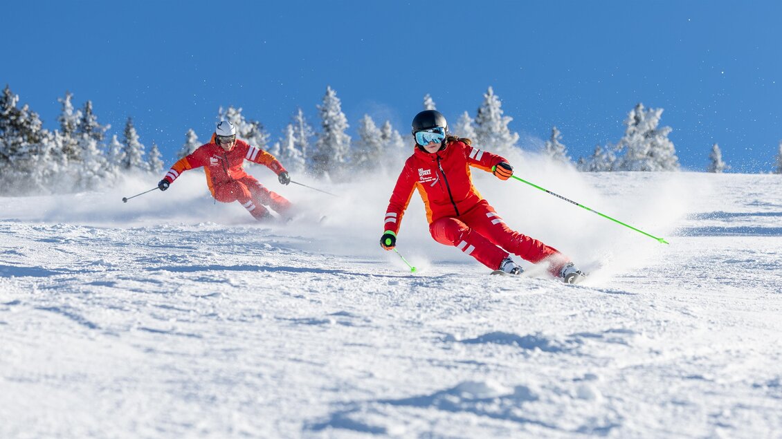 Zwei Skilehrer in roten Anzügen carven dynamisch über die Piste, feiner Schneespray wirbelt auf; im Hintergrund verschneite Bäume und tiefblauer Himmel. | © Skischule Tritscher