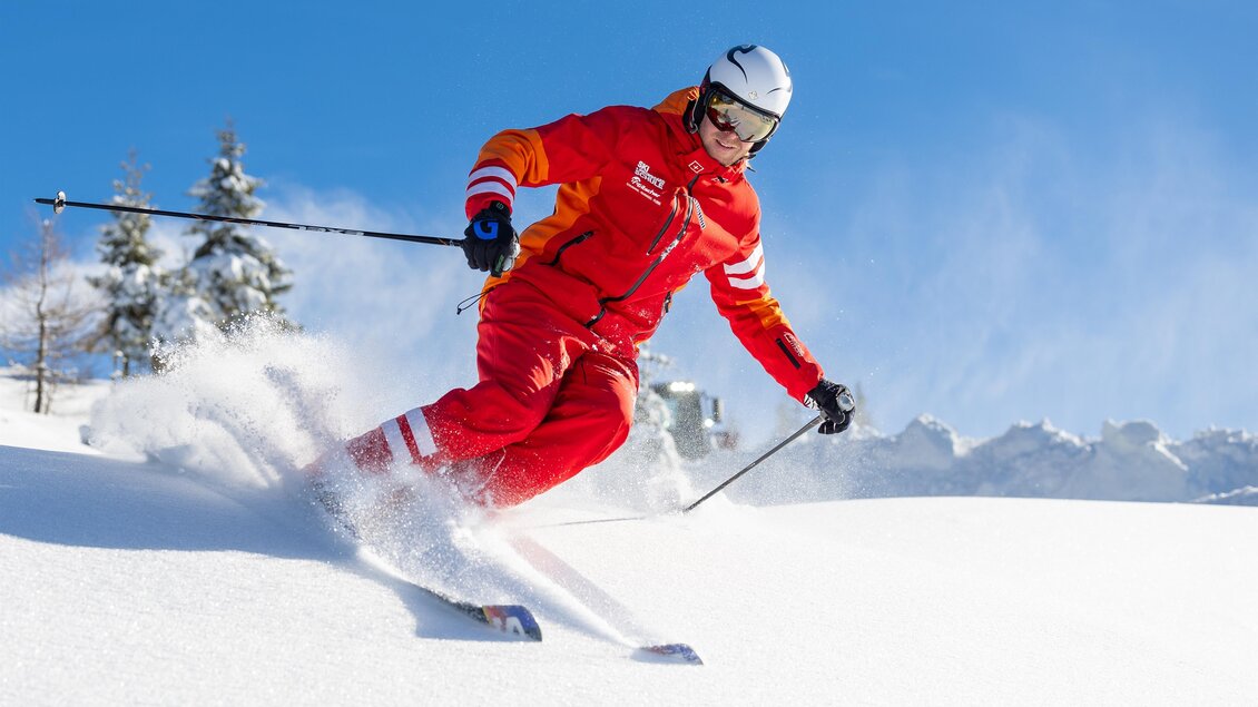 Skilehrer in roter Skischuluniform carvt kraftvoll durch frischen Pulverschnee; Schneestaub fliegt auf, im Hintergrund alpine Winterlandschaft. | © Skischule Tritscher
