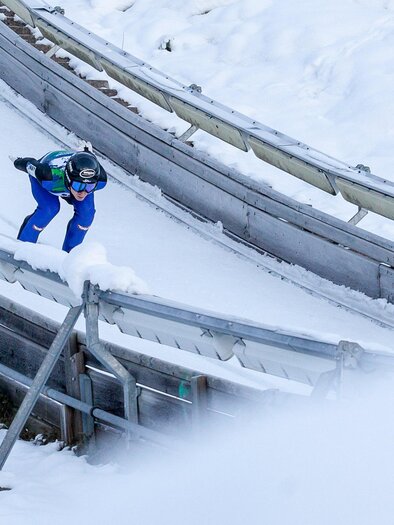 A ski jumper in blue gear is going down the ramp. Snow lies around him and the track is clearly visible. | © ÖSV/ FLORIAN KOTLABA