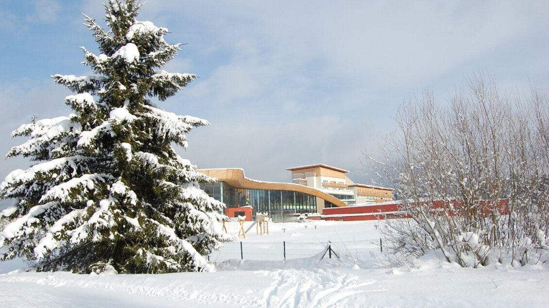 Ein winterliches Landschaftsbild mit schneebedeckten Bäumen und einem modernen Gebäude im Hintergrund. Der Himmel ist klar und blau. | © Aldiana Salzkammergut und GrimmingTherme