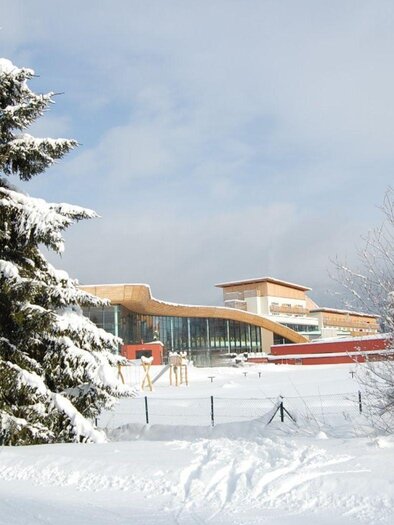A winter landscape with snow-covered trees and a modern building in the background. The sky is clear and blue. | © Aldiana Salzkammergut und GrimmingTherme