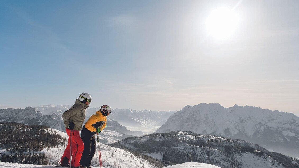 Zwei Skifahrer stehen auf einem schneebedeckten Berg und genießen die Aussicht. Der Himmel ist blau und die Sonne strahlt hell. | © Aldiana Salzkammergut und GrimmingTherme