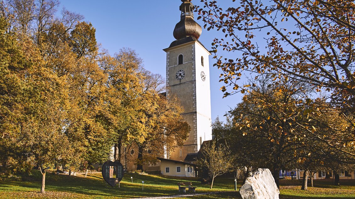 Eine schöne Kirche mit einem hohen Turm steht umgeben von bunten Herbstbäumen. Ein ruhiger Weg führt durch die grüne Wiese. | © Armin Walcher