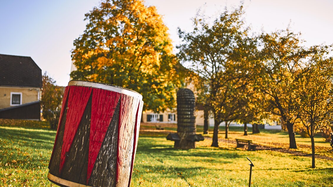 Ein bunter Trommel steht im Vordergrund, umgeben von bunten Bäumen im Herbst. Im Hintergrund sind einige Skulpturen und Bänke in einem ruhigen Park sichtbar. | © Armin Walcher
