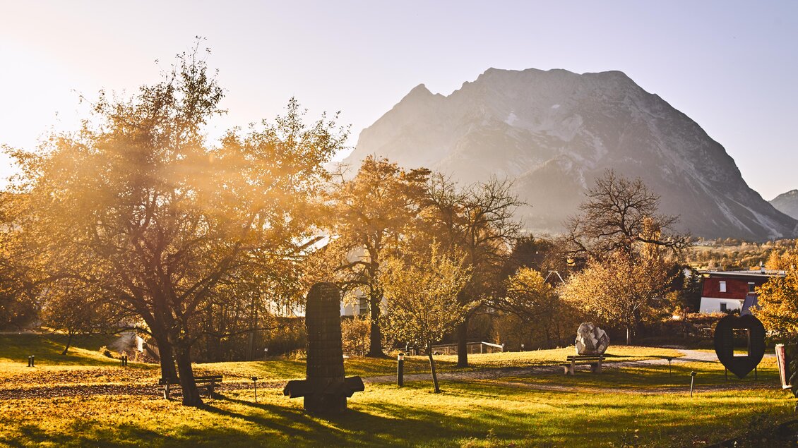 Eine malerische Landschaft mit Bäumen und Skulpturen im Vordergrund. Im Hintergrund erhebt sich ein beeindruckender Berg unter einem schönen Sonnenlicht. | © Armin Walcher