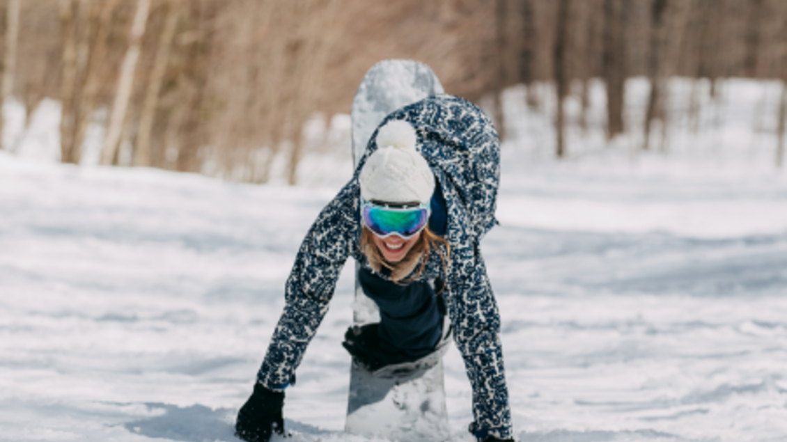 Eine Person mit einem Snowboard sitzt im Schnee und lächelt. Im Hintergrund sind verschneite Bäume sichtbar. | © Burton