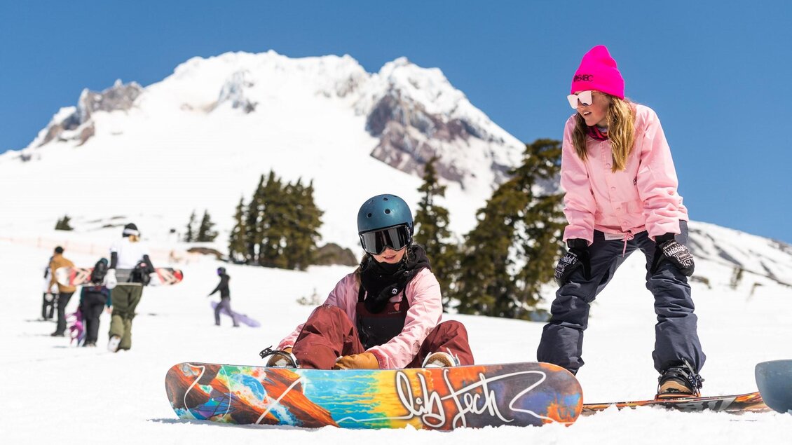Zwei Frauen snowboarden auf verschneitem Gelände. Im Hintergrund sind schneebedeckte Berge und andere Wintersportler zu sehen. | © Lib Tech