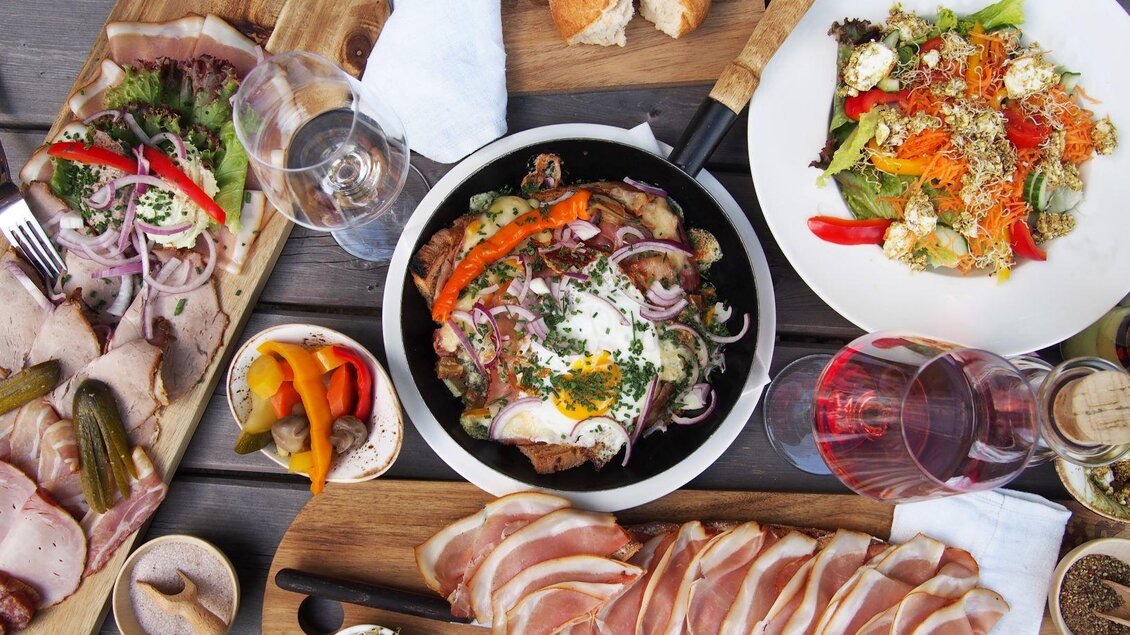 Ein reichhaltiges Essen auf einem Tisch, bestehend aus verschiedenen Fleischplatten, einer bunten Salatschüssel und einer Pfanne mit Gemüse. Dazu werden Gläser mit Wein und Brot serviert. | © Rogner Bad Blumau