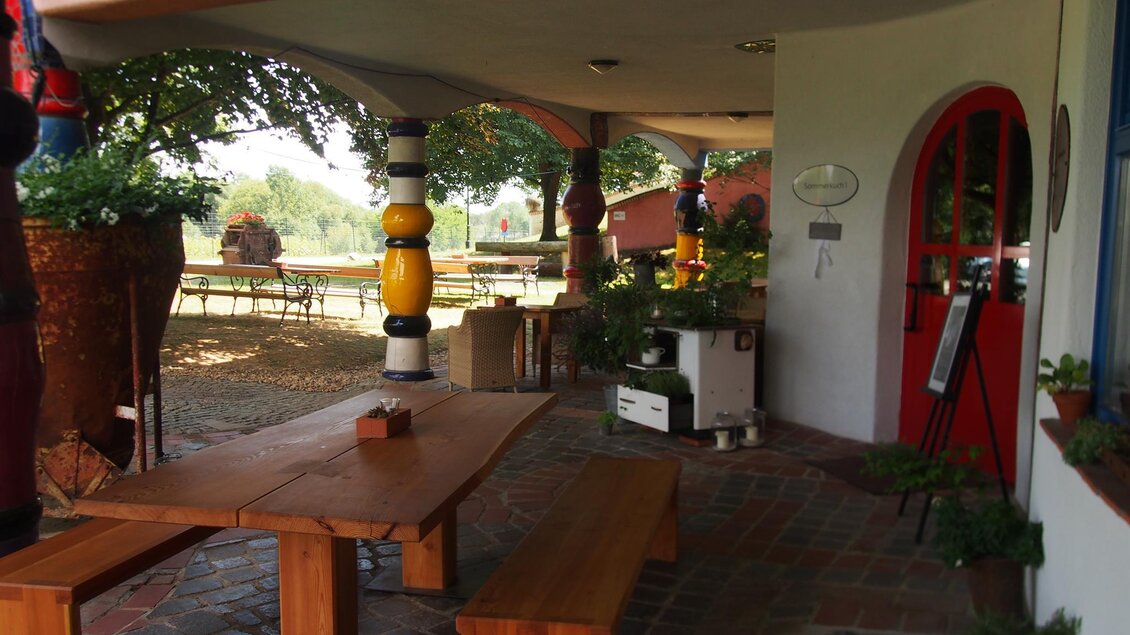 Ein einladender Außenbereich mit einem großen Tisch aus Holz und bunten Säulen. Der Raum wirkt gemütlich und bietet Platz für entspannte Stunden im Freien. | © Rogner Bad Blumau
