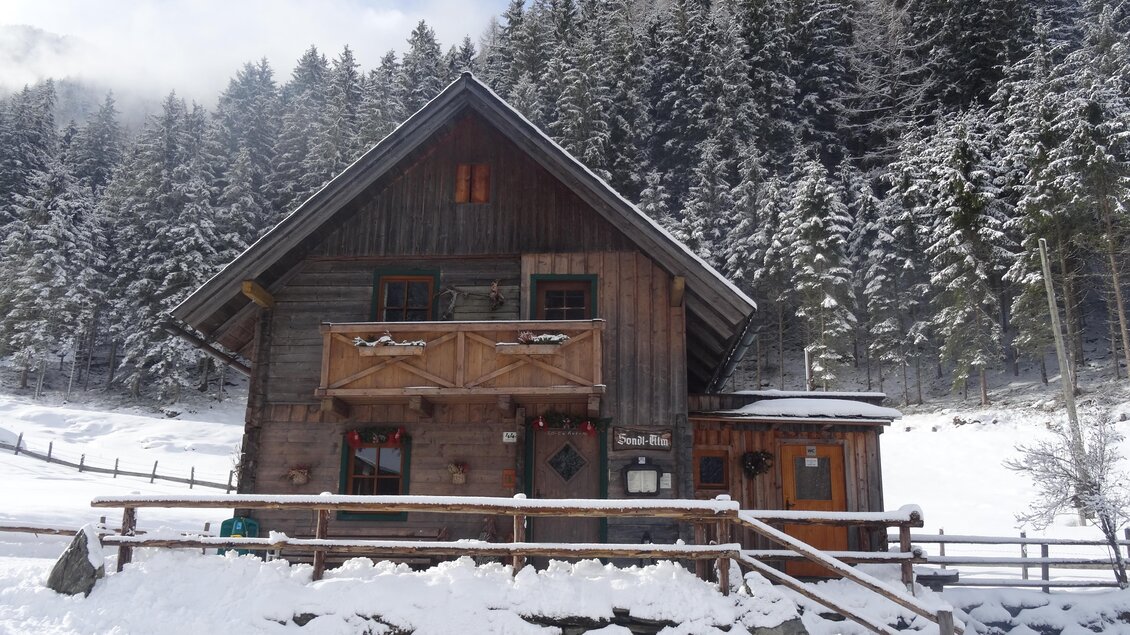 Ein gemütliches Holzhaus in schneebedeckter Landschaft. Im Hintergrund sind hohe, grüne Bäume zu sehen. | © Sondl-Alm