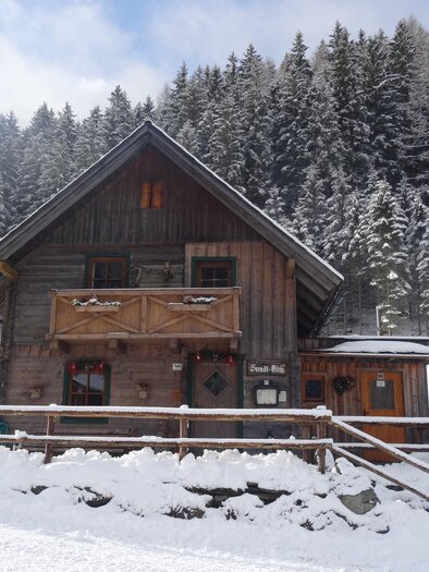 A cozy wooden house in a snow-covered landscape. In the background, tall, green trees can be seen. | © Sondl-Alm
