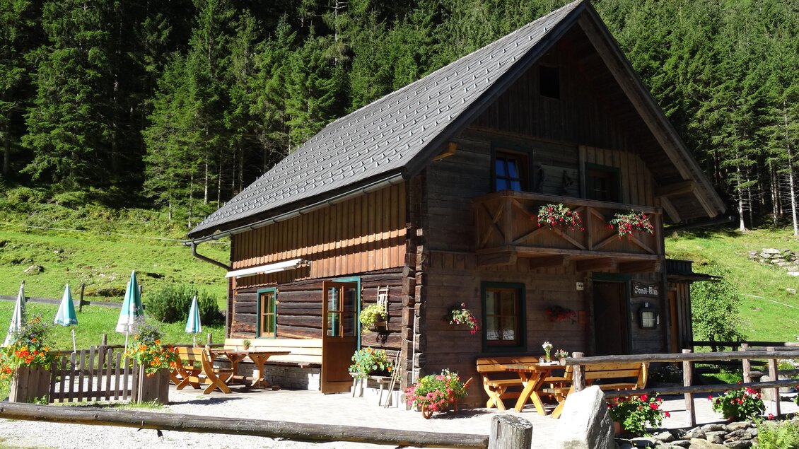 Ein charmantes Holzhaus in der Natur, umgeben von Bäumen und Blumen. Die Terrasse ist mit Tischen und Sonnenschirmen ausgestattet. | © Sondl-Alm
