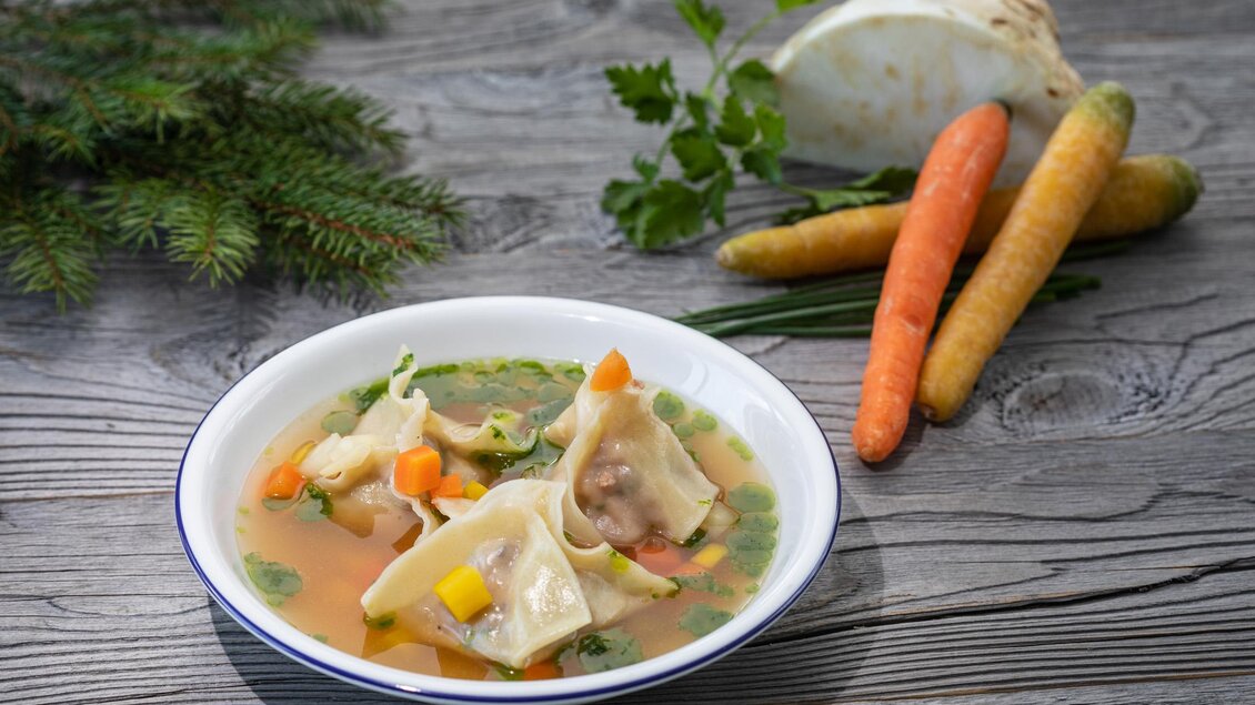Eine Schüssel mit frischer Suppe, gefüllt mit Gemüse und Teigtaschen. Neben der Schüssel liegen einige Karotten und ein Kohlkopf auf einem Holztisch. | © Sondl-Alm
