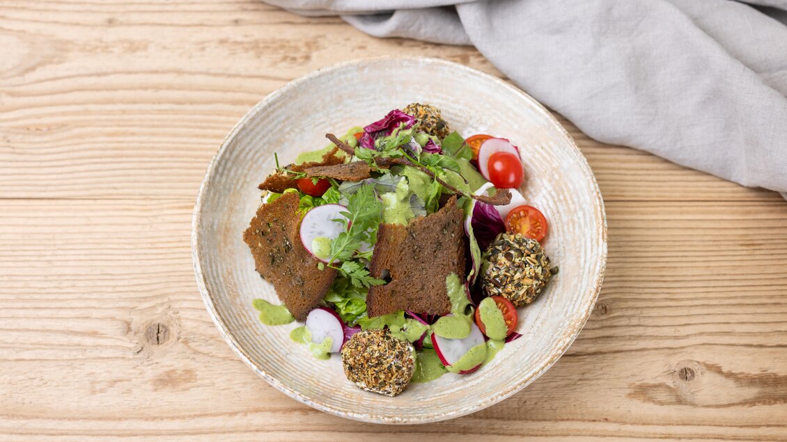Ein frischer Salat mit buntem Gemüse und knusprigen Brotscheiben. Dazu sind würzige Bällchen dekorativ angerichtet. | © Sondl-Alm