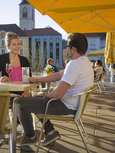 Eine fröhliche Unterhaltung zwischen zwei Personen in einem Café im Freien. Sonnige Atmosphäre mit Plätzen und Sonnenschirmen. | © M.Maili