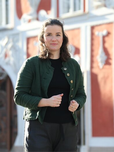 A woman is standing, smiling, in front of a historic building with red walls. She is wearing a green jacket and looks friendly and welcoming. | © TV ERZBERG LEOBEN