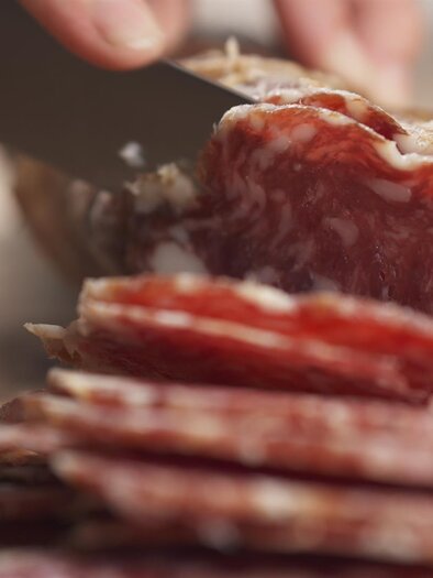 Eine Hand schneidet dünne Scheiben von Salami mit einem Messer. Im Hintergrund ist ein Holzbrett sichtbar. | © GCapture/Shutterstock