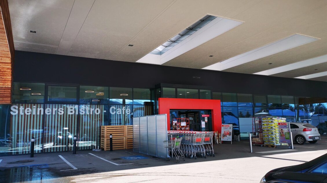 Ein modernes Bistro mit großen Fenstern und einem Eingangsbereich. Parkplätze und Einkaufswagen sind im Vordergrund sichtbar. | © TV Hochsteiermark | Brigitte Digruber