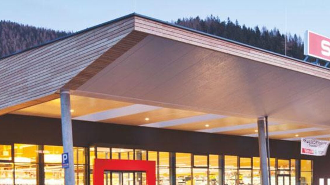 Ein moderner Supermarkt mit Holzfassade und großem Dach. Im Hintergrund sind Berge zu sehen, und die Beleuchtung des Geschäfts wirkt einladend. | © www.sparsteiner.at