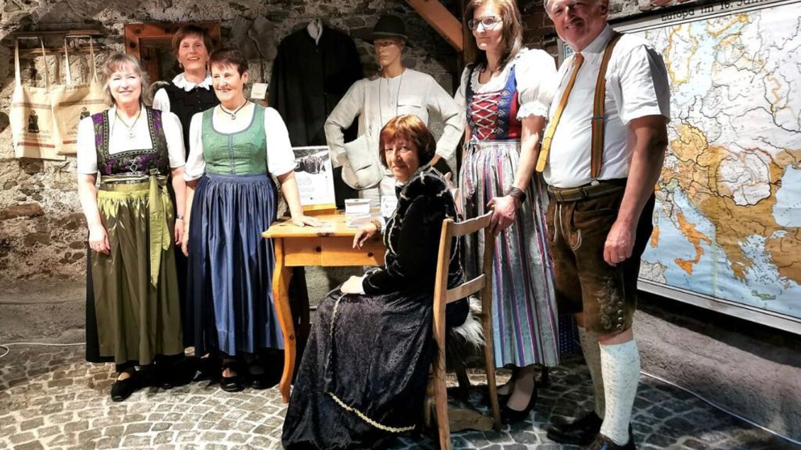 Eine Gruppe von Menschen in traditioneller Tracht steht in einem Museum. Im Hintergrund sind historische Kleider und eine Karte zu sehen. | © TVB Murau