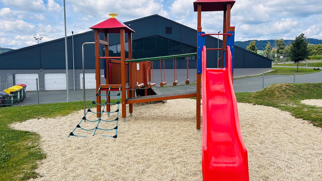 Ein Spielplatz mit einer roten Rutsche, einer Kletterwand und Schaukeln. Der Platz ist mit Sand bedeckt und hat eine grüne Umgebung. | © Tourismus Vorau