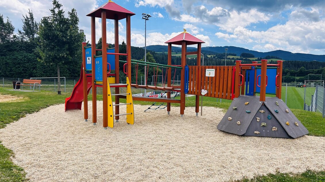 Ein farbenfroher Spielplatz mit Rutschen und einem Klettergerüst steht in einer grünen Umgebung. Im Hintergrund sind Hügel und ein Sportplatz zu sehen. | © Tourismus Vorau