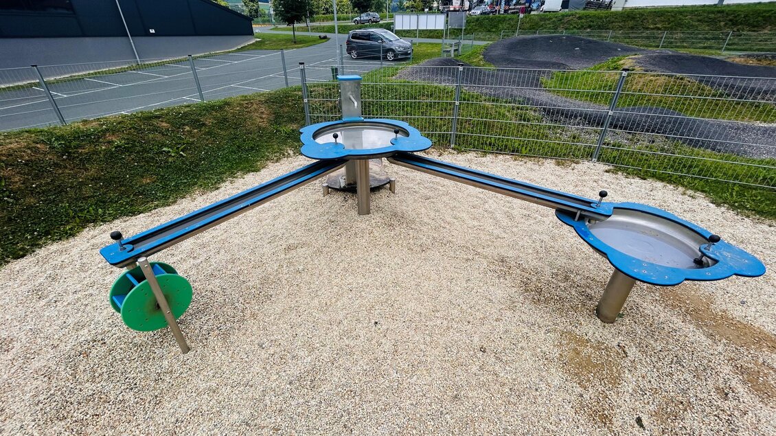 Ein Spielplatzgerät mit drei Schalen, die an einem zentralen Punkt verbunden sind. Die Oberfläche besteht aus Kies, und im Hintergrund sind Gebäude und eine Straße zu sehen. | © Tourismus Vorau