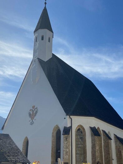 Eine weiße Kirche mit einem spitzen Turm steht unter einem klaren blauen Himmel. Im Hintergrund sind Berge und mehrere Gebäude zu sehen. | © TVB Ausseerland Salzkammergut/Jana Grill