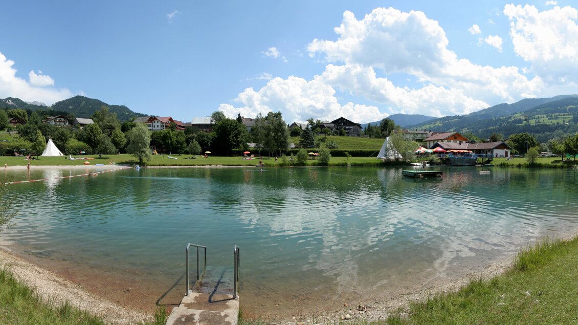 Ein ruhiger See umgeben von grünen Wiesen und Bergen. Der Himmel ist klar mit einigen Wolken, und es gibt Boote im Wasser. | © Bärenwirt