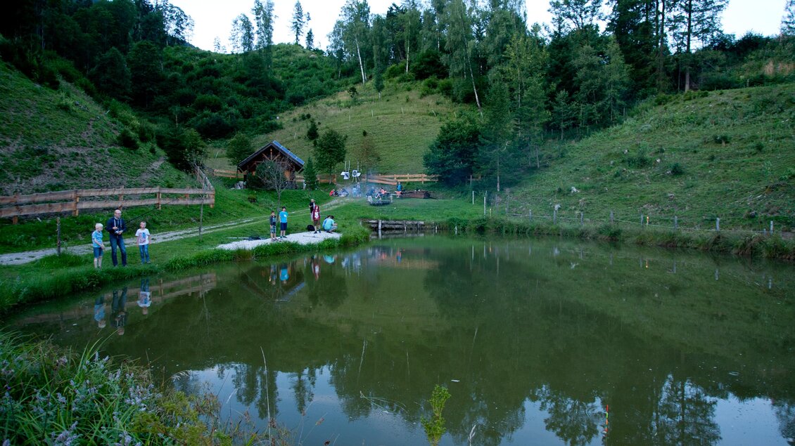 Ein ruhiger Teich umgeben von grünen Hügeln und Bäumen. Menschen genießen die ländliche Atmosphäre in der Natur. | © Bärenwirt