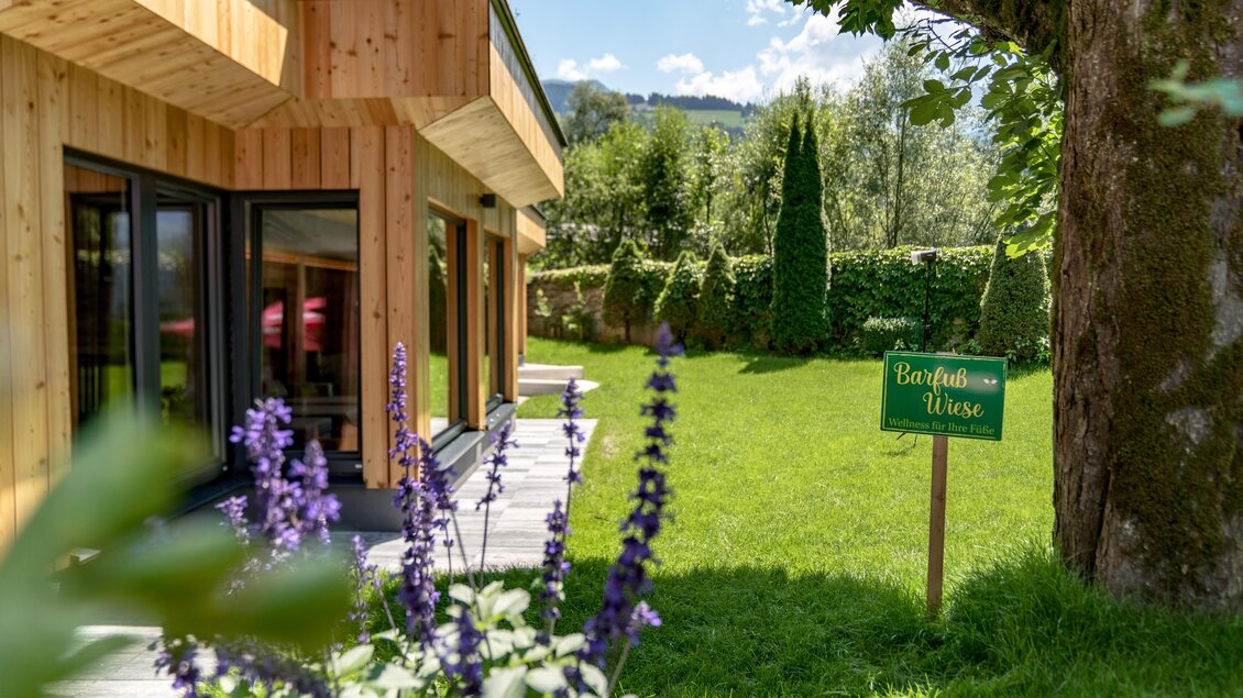 Ein modernes Haus mit Holzfassade und großen Fenstern steht in einem gepflegten Garten. Im Vordergrund blühen lila Blumen und ein Schild mit der Aufschrift „Barfuß Wiese“ ist sichtbar. | © baerenwirt.com