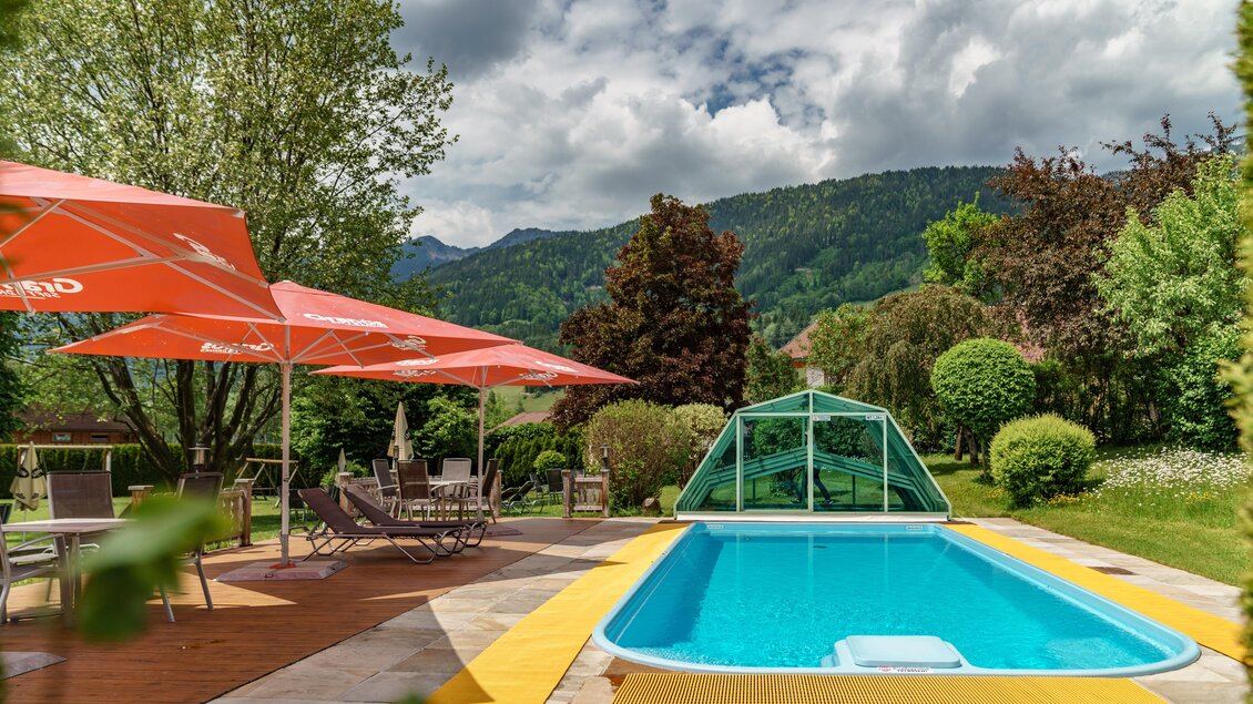 Ein schöner Pool in einem Garten mit grünen Bäumen und Bergen im Hintergrund. Sonnenschirme schützen die Liegen vor der Sonne. | © baerenwirt.com