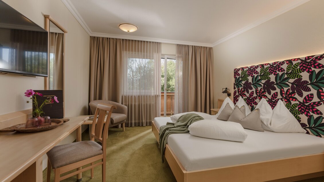 Ein modernes Hotelzimmer mit zwei Betten und buntem Kopfteil. Ein Tisch, Stuhl und ein kleiner Balkon mit Blick auf die Natur ergänzen den Raum. | © baerenwirt.com