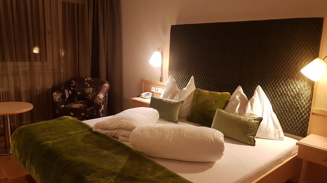Ein gemütliches Schlafzimmer mit einem großen Bett, das mit grünen und weißen Kissen dekoriert ist. Neben dem Bett steht ein Sessel und es gibt sanfte Beleuchtung. | © Bärenwirt