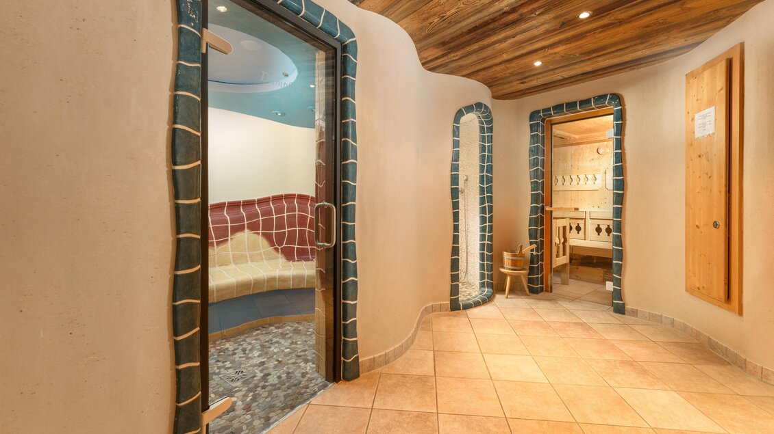 Ein entspannender Wellnessbereich mit geschwungenen Wänden und mehreren Zugängen zu unterschiedlichen Räumen. Der Boden ist gefliest und die Decke hat ein warmes Holzdesign. | © baerenwirt.com