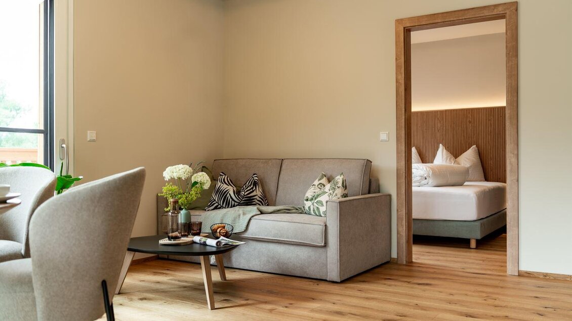Helles, gemütliches Wohnzimmer mit grauem Sofa, dekorativen Kissen und Holzboden, daneben ein Durchgang zu einem Schlafzimmer mit Doppelbett und Holzrückwand. | © Bärenwirt