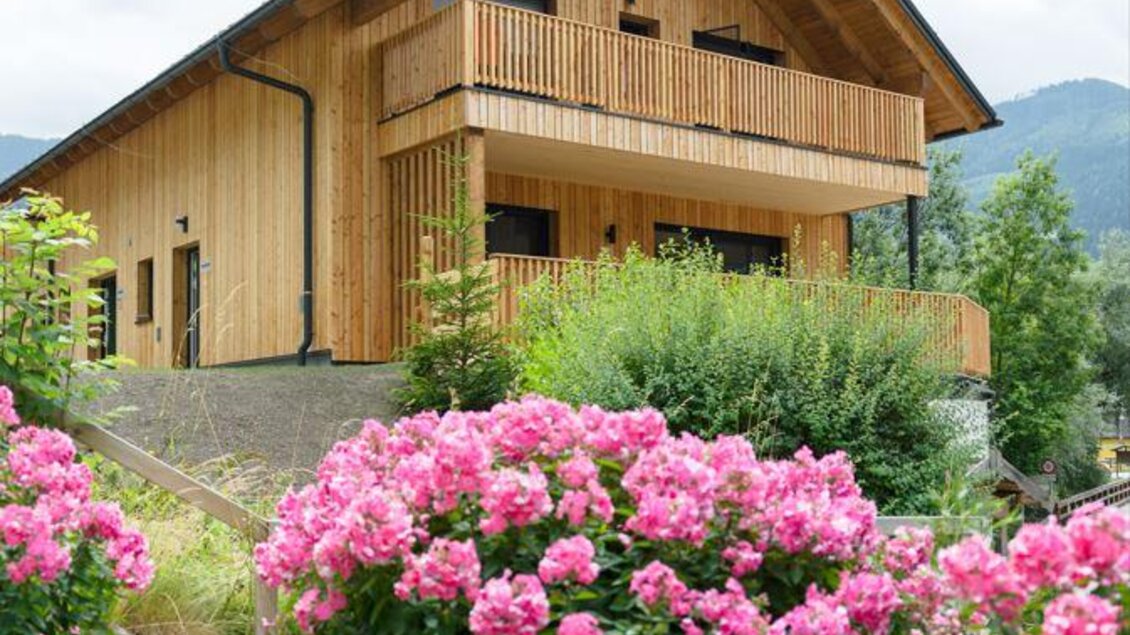 Modernes Chalet aus hellem Holz mit großem Balkon, umgeben von einem gepflegten Garten mit bunten Blumenbeeten und Berglandschaft im Hintergrund. | © Bärenwirt