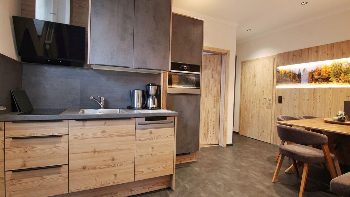 Gemütliche Appartementküche mit Holzfronten und dunklen Oberflächen, komplett ausgestattet, daneben ein Esstisch mit Polsterstühlen und stimmungsvollem Wandbild. | © Bärenwirt