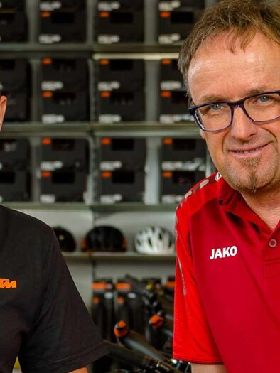 Two men are smiling in a bicycle shop. Bicycles and clothing are visible in the background. | © Josef Rabensteiner