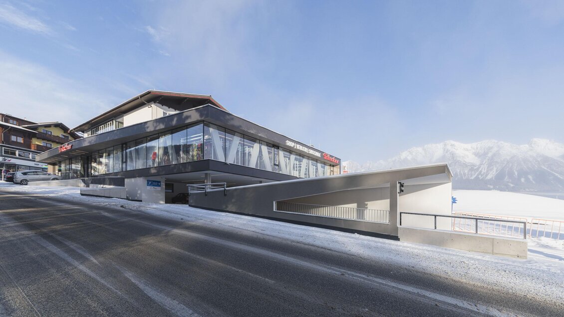 Ein modernes Gebäude mit großen Glasfenstern in einer schneebedeckten Umgebung. Im Hintergrund sind Berge unter einem klaren Himmel zu sehen.