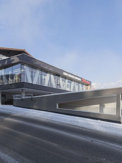 A modern building with large glass windows in a snow-covered setting. In the background, mountains can be seen under a clear sky.