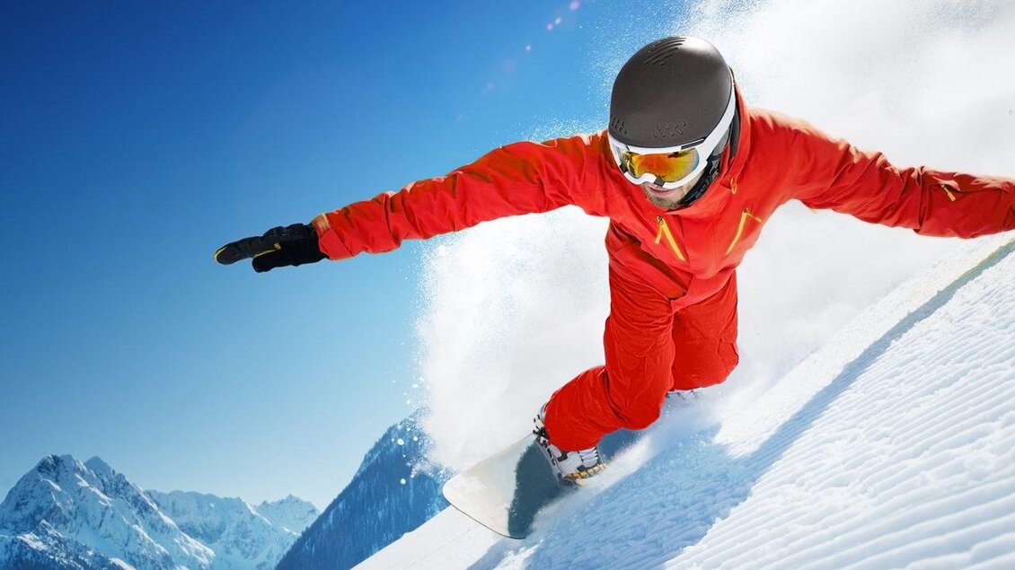Ein Snowboarder in einem roten Anzug fährt eine schneebedeckte Piste hinunter. Im Hintergrund sind schneebedeckte Berge und ein blauer Himmel zu sehen. | © Skischule Tritscher