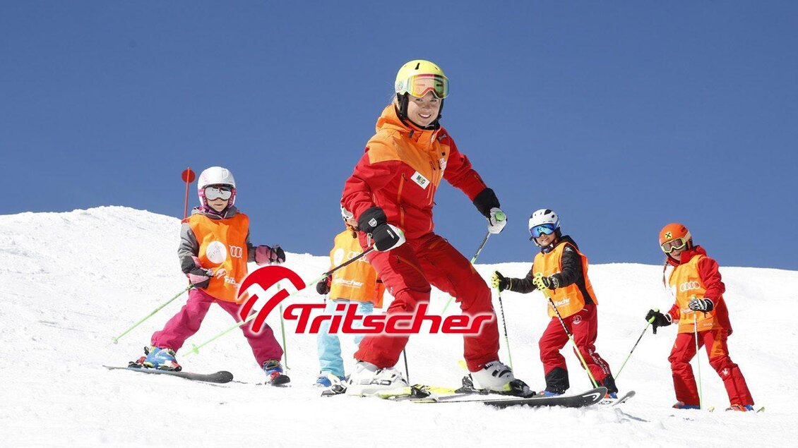 Eine Gruppe von Kindern, die Ski fahren, wird von einem Lehrer angeleitet. Sie tragen bunte Skikleidung und sind auf einer verschneiten Piste. | © Skischule Tritscher