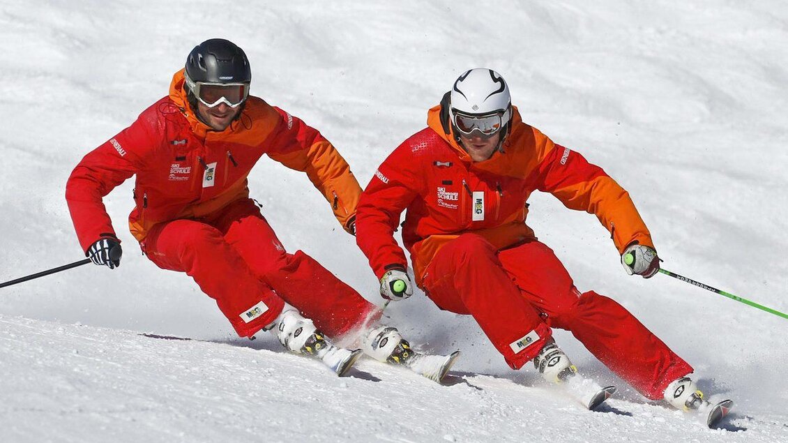 Zwei Skifahrer in roten Anzügen fahren nebeneinander auf einer schneebedeckten Piste. Die Sonne scheint und der Schnee glitzert. | © Skischule Tritscher