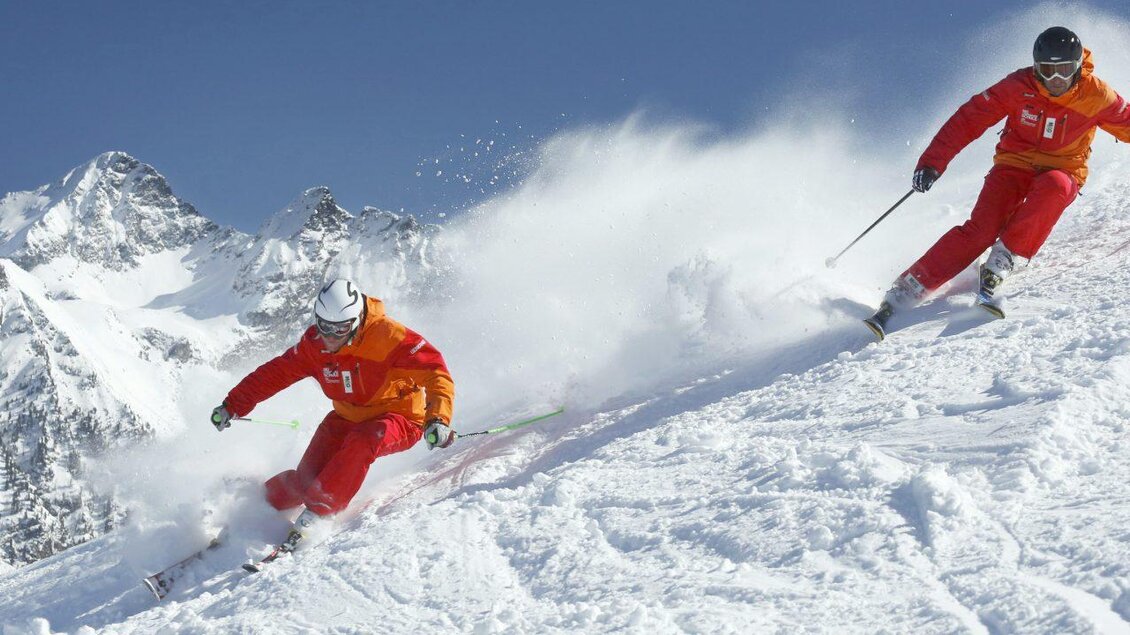 Zwei Skifahrer in roten Anzügen fahren auf einer verschneiten Piste. Im Hintergrund sind hohe Berggipfel und ein klarer blauer Himmel zu sehen. | © Skischule Tritscher