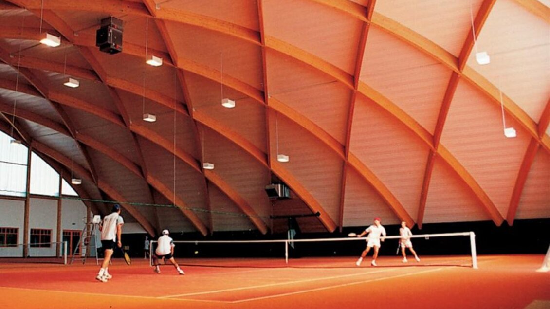 Eine Sporthalle mit einem hohen, gewölbten Dach. Im Vordergrund spielen mehrere Personen Tennis auf einem roten Platz. | © Lederer
