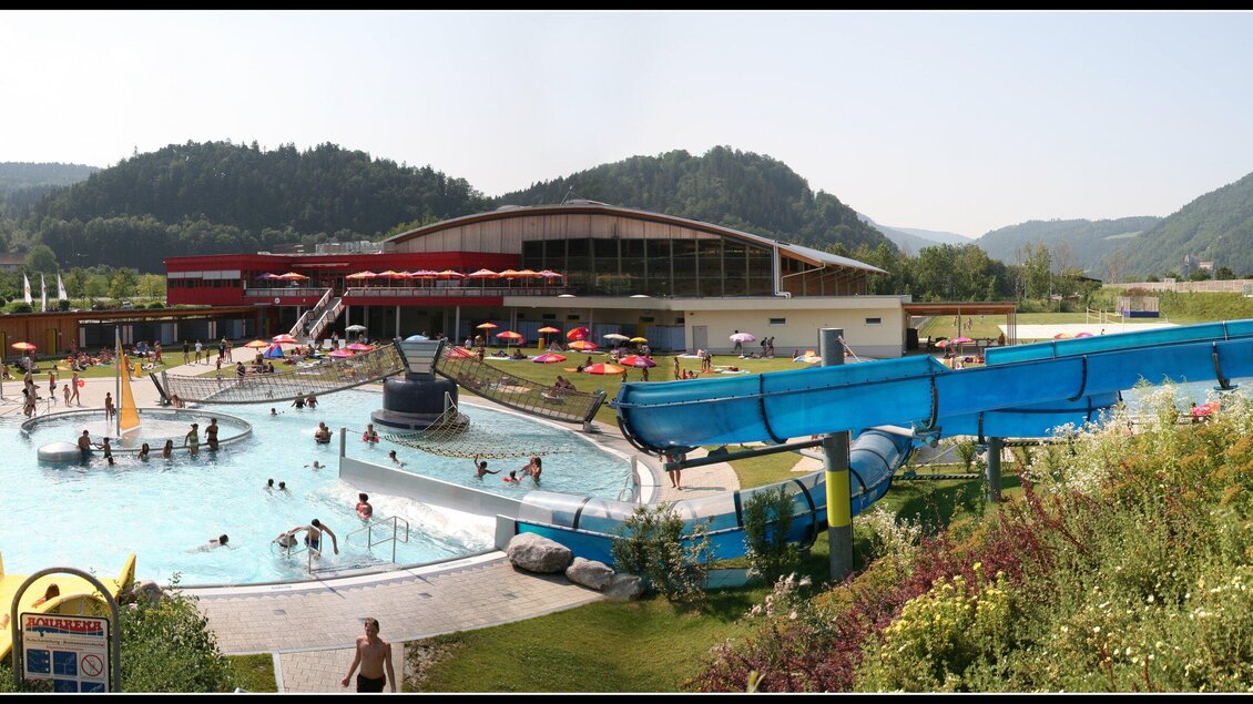 Der Freizeitpark Frohnleiten mit verschiedenen Rutschen und einem Schwimmbecken. Im Hintergrund sind Berge und Sitzbereiche mit Sonnenschirmen zu sehen, sowie die Eishalle. | © TV Region Graz - Helmut Lunghammer